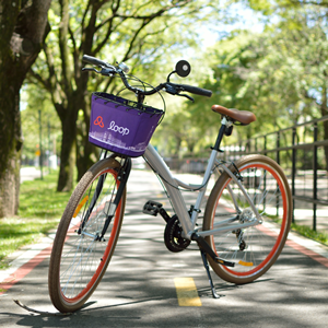 Bicicleta da Loop em uma ciclovia, com fundo repleto de árvores. A bike está posicionada de forma que é possível ver toda a bike: a cestinha roxa em que está gravado o nome da empresa 'Loop', o corpo da bike em cinza e e as duas rodas marrom com detalhes laranjas.