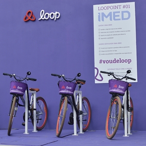 Imagem do primeiro Loopoint disponível, localizado na IMED Porto Alegre. Na fotografia há três bicicletas da Loop presas aos seus respectivos paraciclos. Ao fundo, há a parede roxa personalizada do Loopoint com a placa que contém uma explicação do modo como o usuário deve retirar e devolver a sua bicicleta. 
